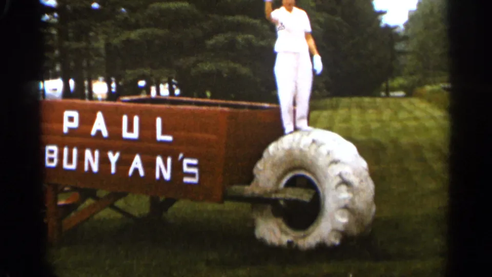 A woman in white stands on the large wheel next to the wooden Paul Bunyan's wheelbarrow display in Duluth, Minnesota, 1965.