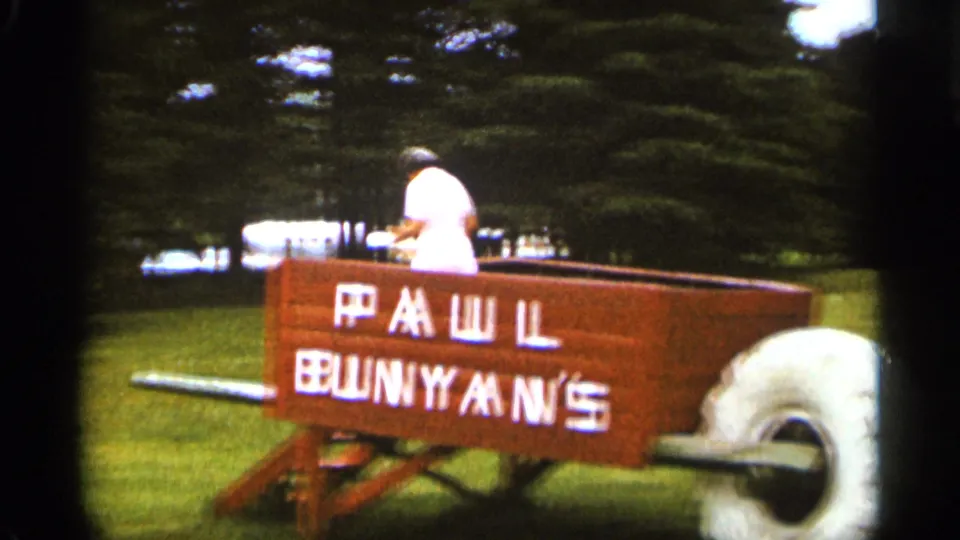 The woman is partly turned away near the wooden Paul Bunyan's wheelbarrow display, with trees and grass behind her in the 1965 Duluth footage.