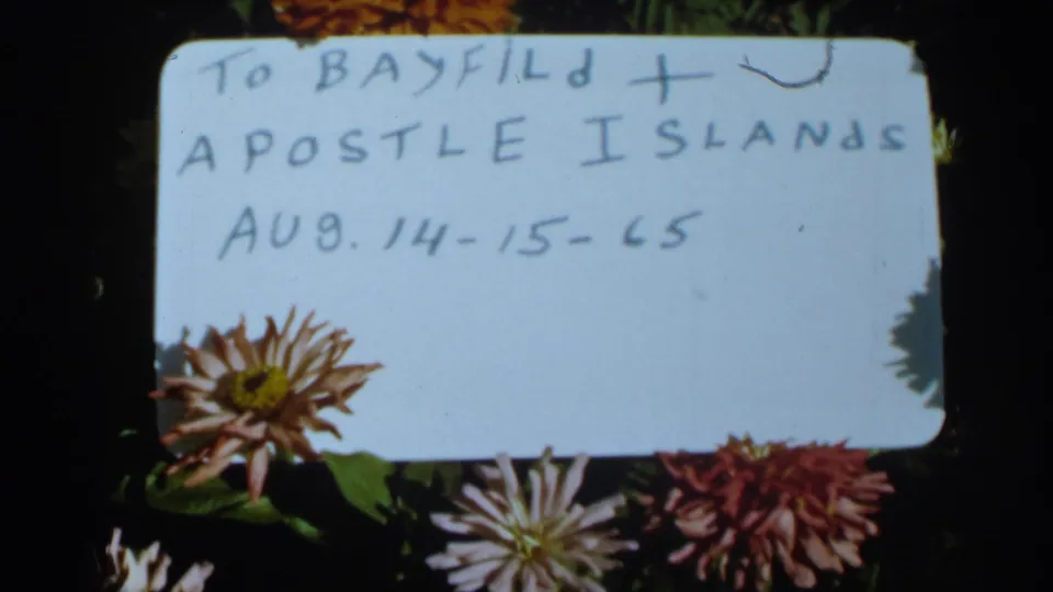 Handwritten 1965 travel card reading Bayfield and Apostle Islands, placed among chrysanthemums on a dark background.