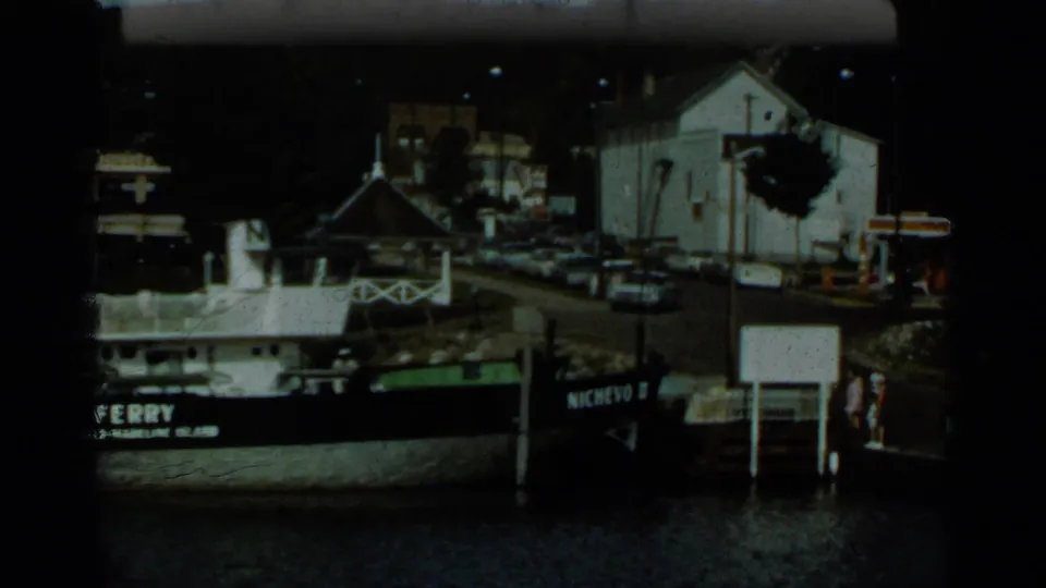 1965 Duluth-area home-movie frame showing a moored ferry at a small waterfront dock with buildings, cars, trees, and water visible.