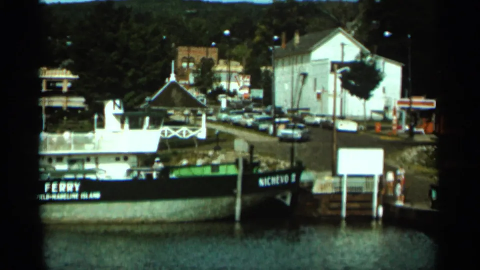 1965 Duluth waterfront frame with the ferry NICHEVO II moored near a pier, backed by parked cars, a white building, and trees.
