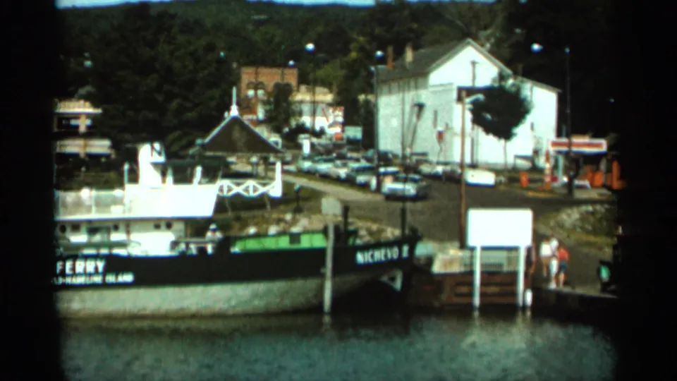 1965 Duluth home-movie frame showing a ferry at the dock, people near the landing, parked cars, and waterfront buildings behind it.