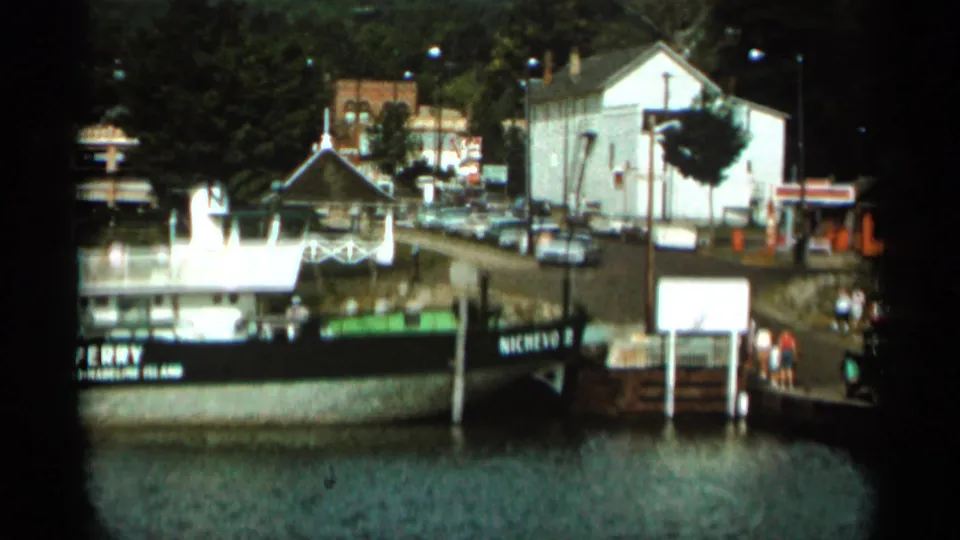 1965 Duluth-area frame of a moored ferry and gangway, with people at the pier, parked cars, trees, and shoreline buildings in view.