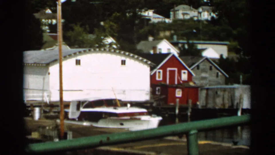1965 Duluth 8mm frame showing a white boathouse, docked boat, red shed, shoreline buildings, and green railing.