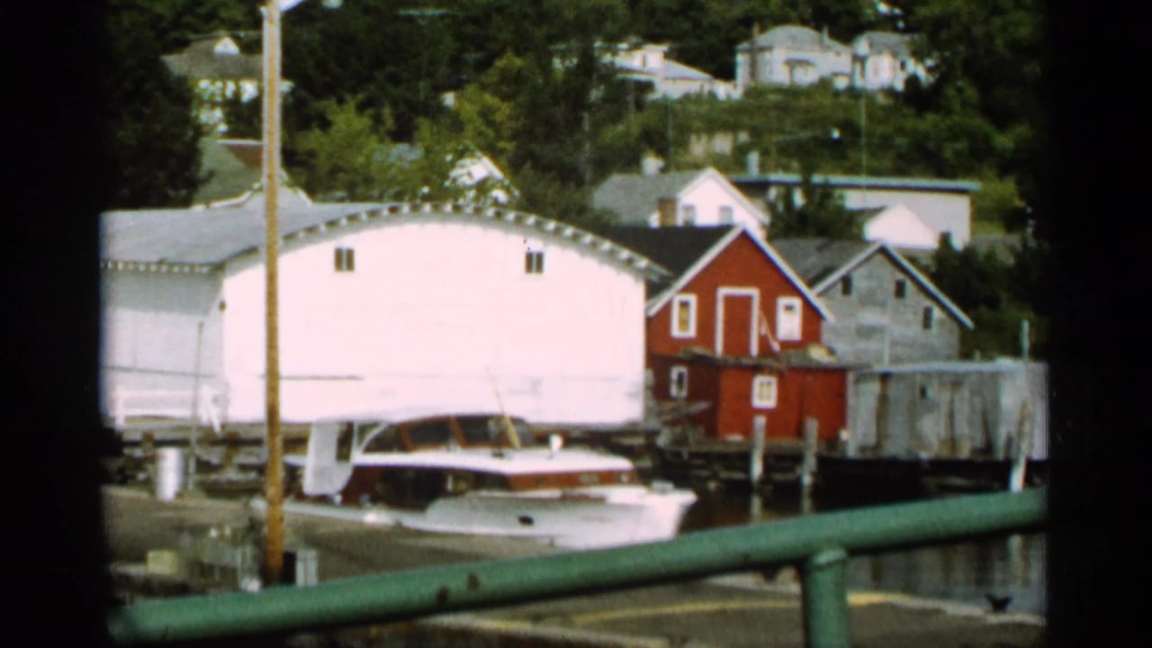 Docked boat near a white boathouse with a red shed and shoreline houses behind.