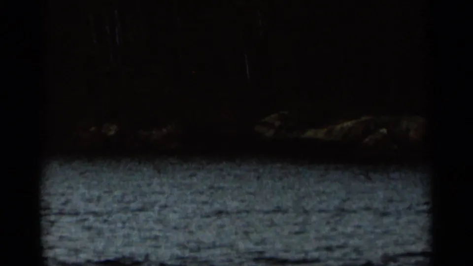 Underexposed 1965 Duluth home-movie frame with rippled water, dark wooded shore, and shoreline rocks.
