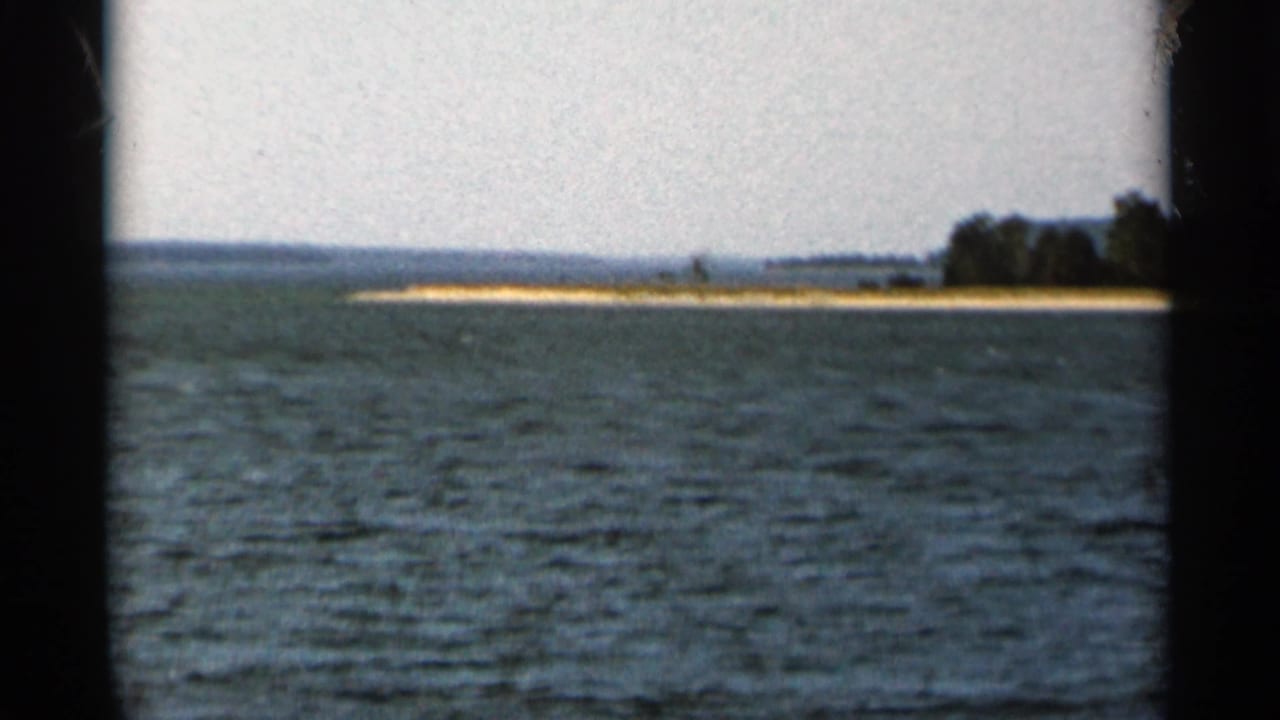 Rippling lake water seen from a boat with a sandy, tree-lined island in the distance.