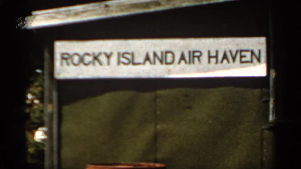 Final handheld frame of the Rocky Island Air Haven sign and green panels, with visible 8mm grain and dark frame edges.