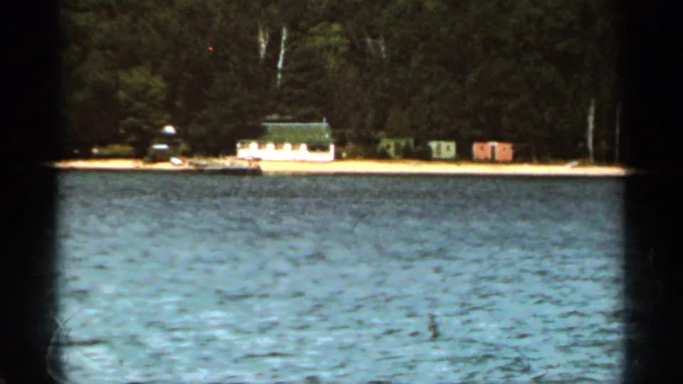 1965 Duluth, Minnesota 8mm frame showing rippled lake water, a wooded shore, small cabins, and a dock in the distance.