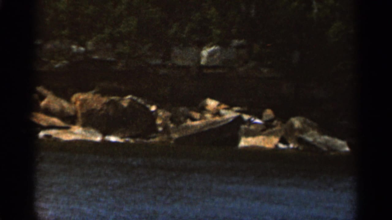 Dark 8mm frame of water, rocks, and a wooded shoreline.