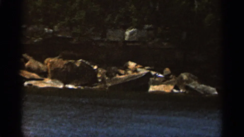1965 Duluth 8mm frame showing rippled water below rocks and dense trees along a shaded shoreline.