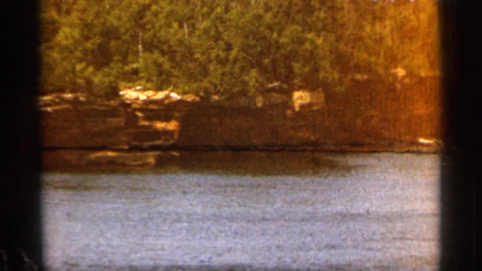 1965 Duluth 8mm frame showing a broad water surface with trees and pale rocks along the far bank.