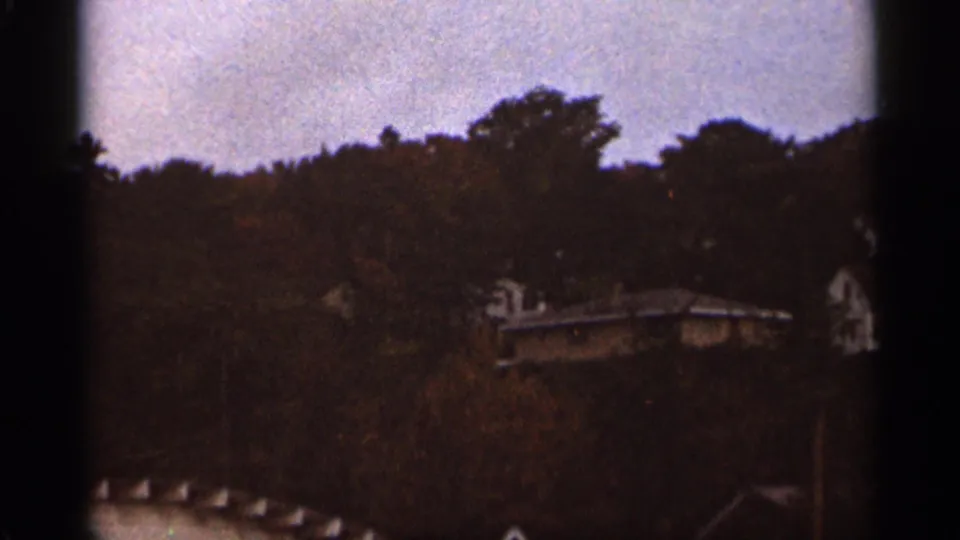 1965 Duluth home-movie frame showing distant house roofs partly hidden by trees beneath a gray sky.