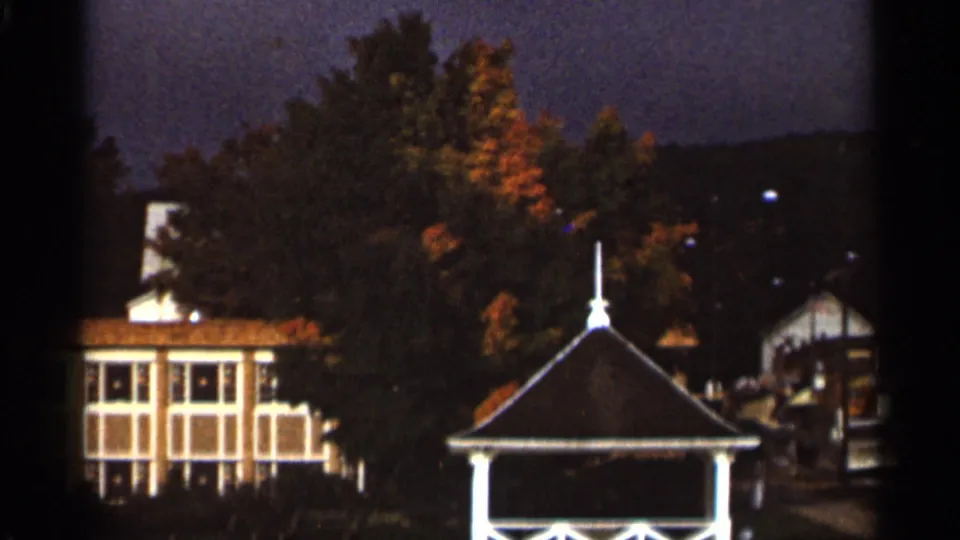 1965 8mm frame from the Duluth metadata showing a gazebo, roadside buildings, and fall foliage in a small-town travel view.