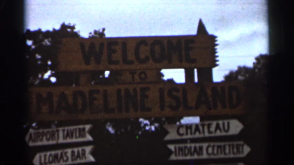 1965 8mm frame showing a wooden Welcome to Madeline Island sign with arrows to local destinations, captured in a grainy travel shot.