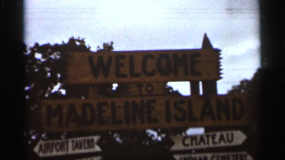 1965 8mm frame showing the Madeline Island welcome sign, partially legible arrow signs, dark trees, and pale sky.