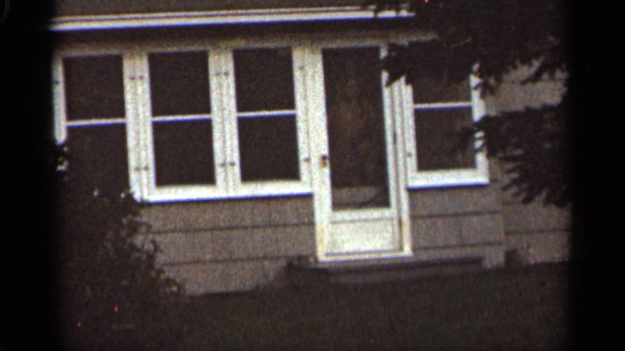 Front of a house with white-trimmed windows and a central door partly framed by dark foliage.