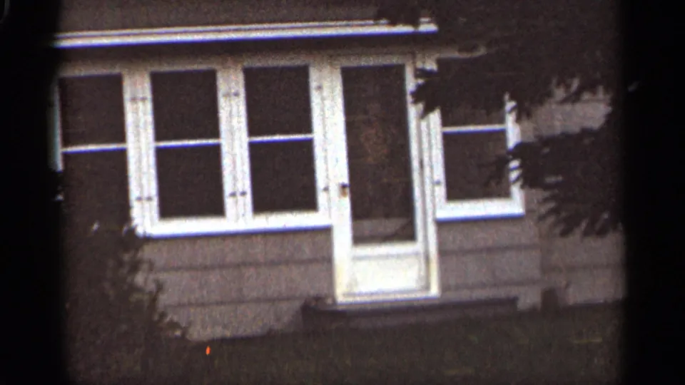 Grainy 1965 Duluth house exterior frame showing white-trimmed windows, a central entry, lawn, and heavy shadow from nearby foliage.