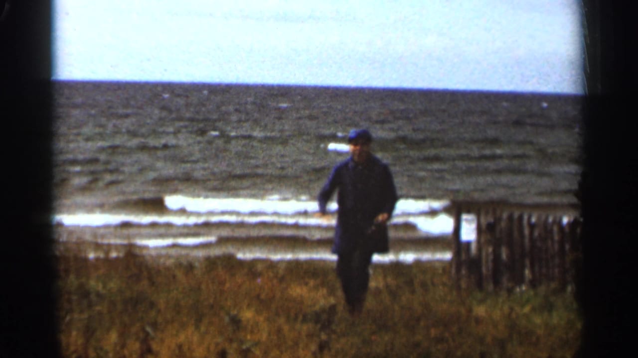 Adult man in a dark coat and cap walks along the Duluth lakeshore, with Lake Superior waves and a wooden fence in the background.