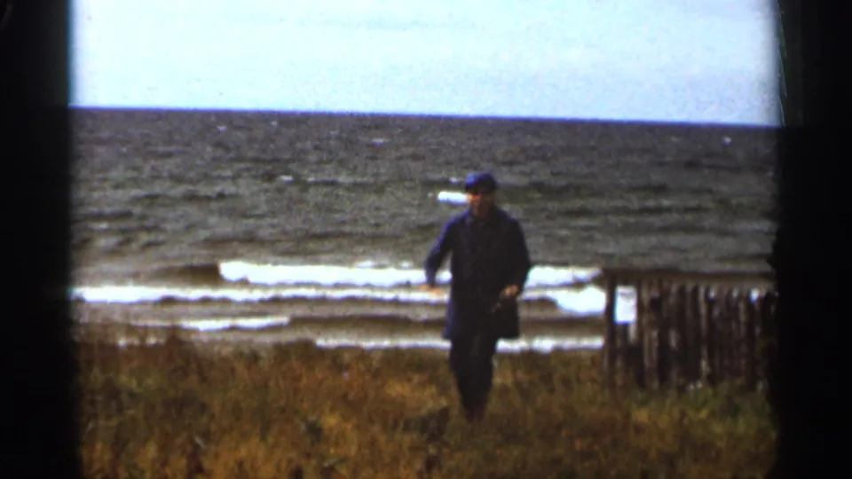 Adult man in a dark coat and cap walks along the Duluth lakeshore, with Lake Superior waves and a wooden fence in the background.