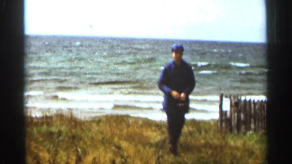 1965 Duluth lakeshore frame with a man in dark clothing facing the camera, grass in the foreground, and choppy Lake Superior water behind him.
