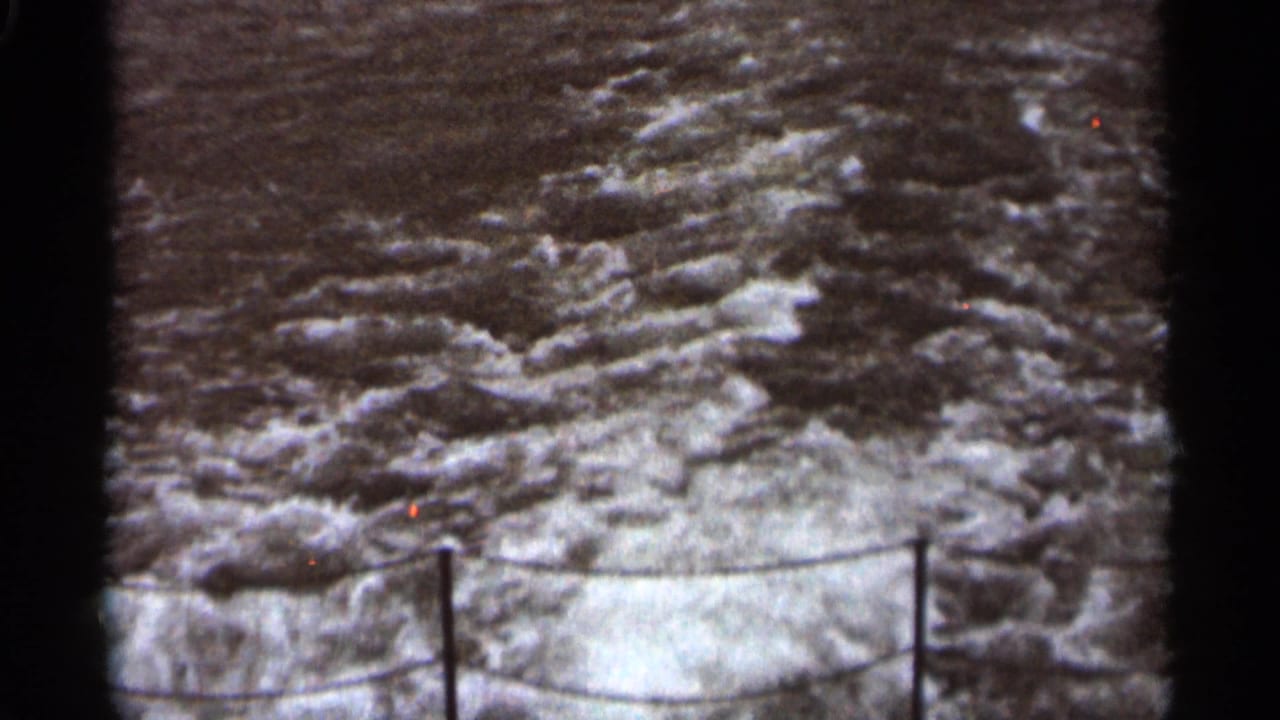 Rough water and whitecaps framed by dark film edges and a low rope rail.