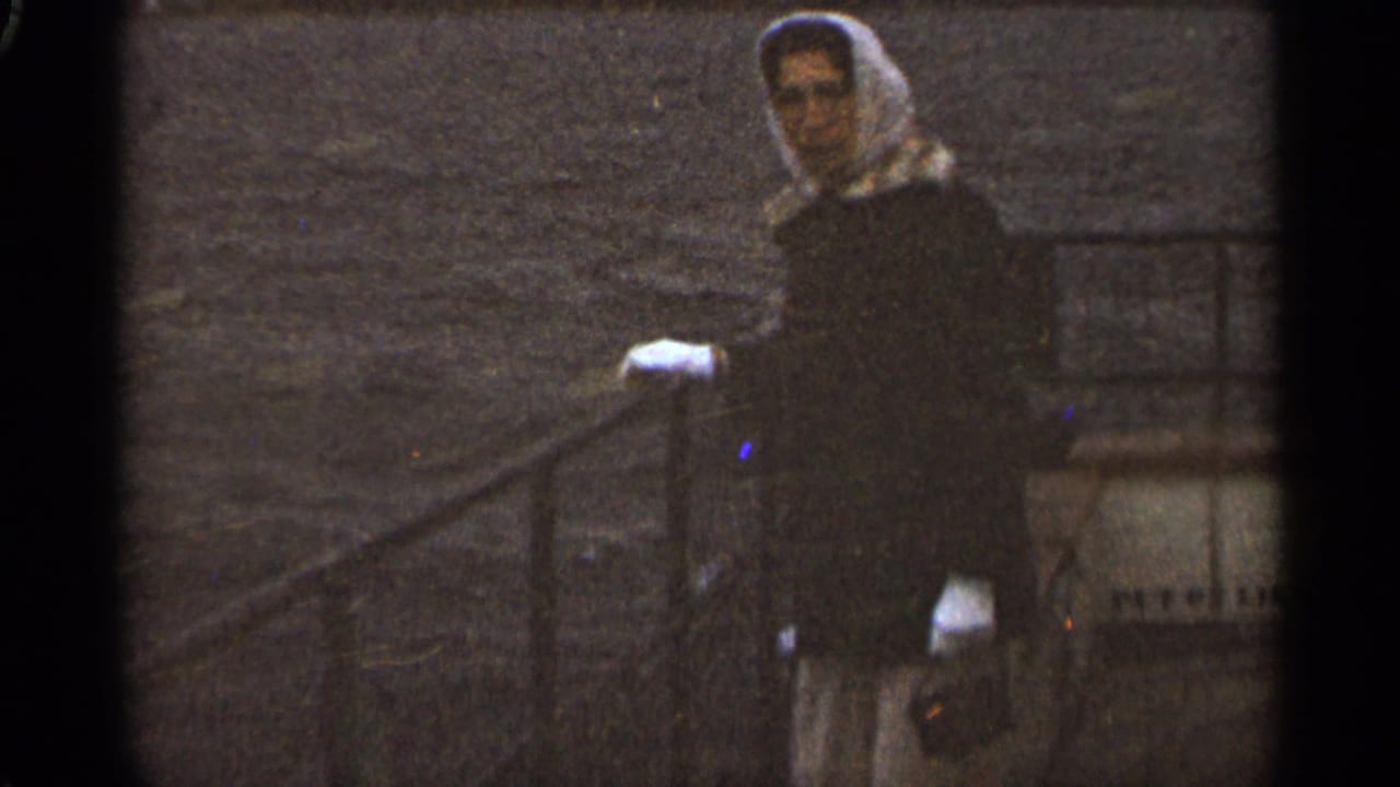 Woman in a white headscarf and dark coat stands by a metal railing on a boat.