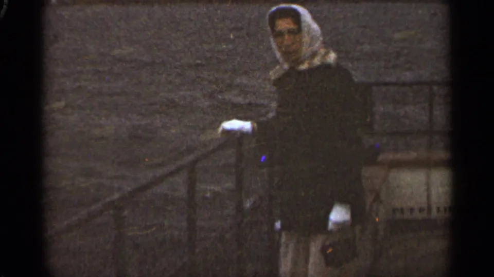 In 1965 Duluth footage, a woman in a white headscarf and dark coat faces the camera near a boat railing with water behind her.