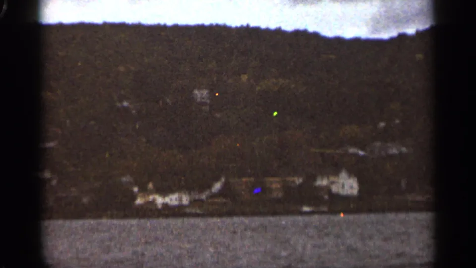 1965 Duluth shoreline view across water, with scattered light-colored buildings at the base of a wooded slope.