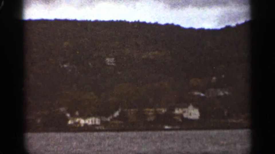 Final 1965 Duluth frame showing a quiet lakeside panorama with water, shoreline buildings, and a dark wooded treeline.