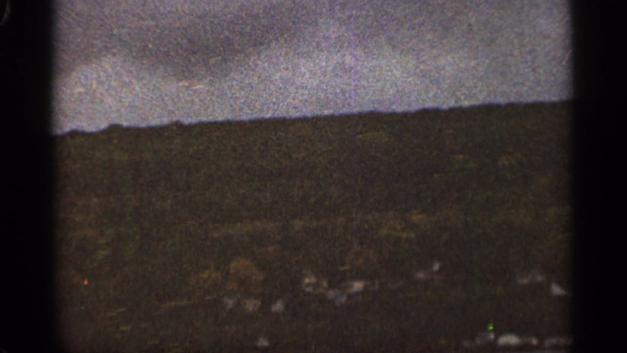 Cloudy sky over a dark wooded hillside with faint distant structures below.