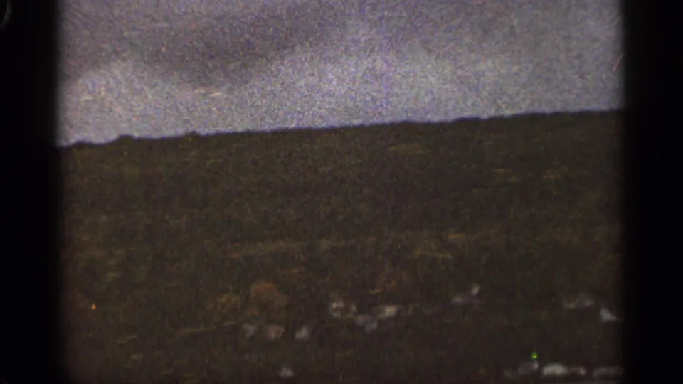 Grainy 1965 Duluth home-movie frame showing a dark hillside under a cloudy sky with small pale structures in the distance.