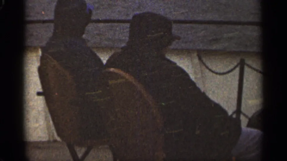 1965 Duluth home-movie frame of two seated silhouetted figures beside a railing, with water or bright ground behind them.