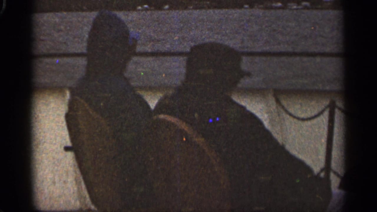 Two dark silhouettes seated side-by-side on a boat near water and shoreline.