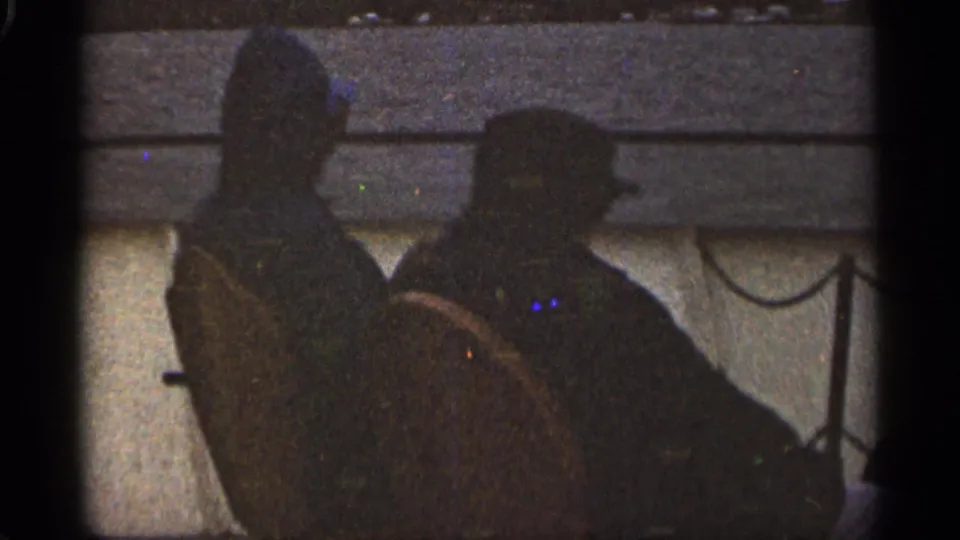 Two adult men appear in silhouette on a boat in supported 1965 Duluth-area 8mm footage, with water and shoreline behind them.