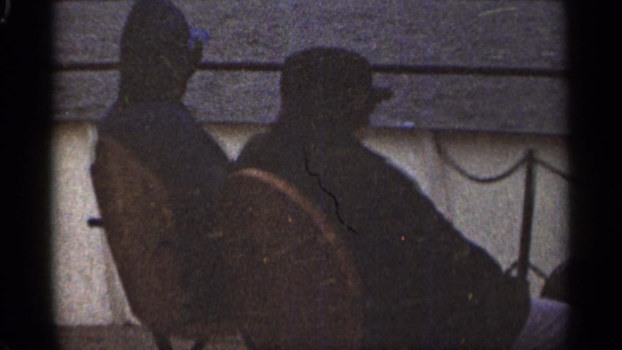 The two men remain seated side-by-side on the boat, seen as dark profiles against water and a distant shore in 1965 Duluth footage.