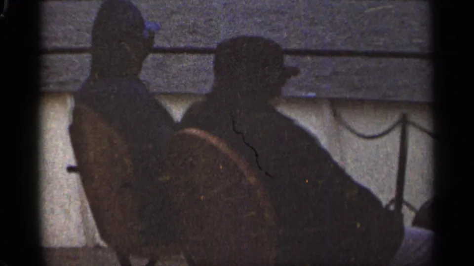 The two men remain seated side-by-side on the boat, seen as dark profiles against water and a distant shore in 1965 Duluth footage.