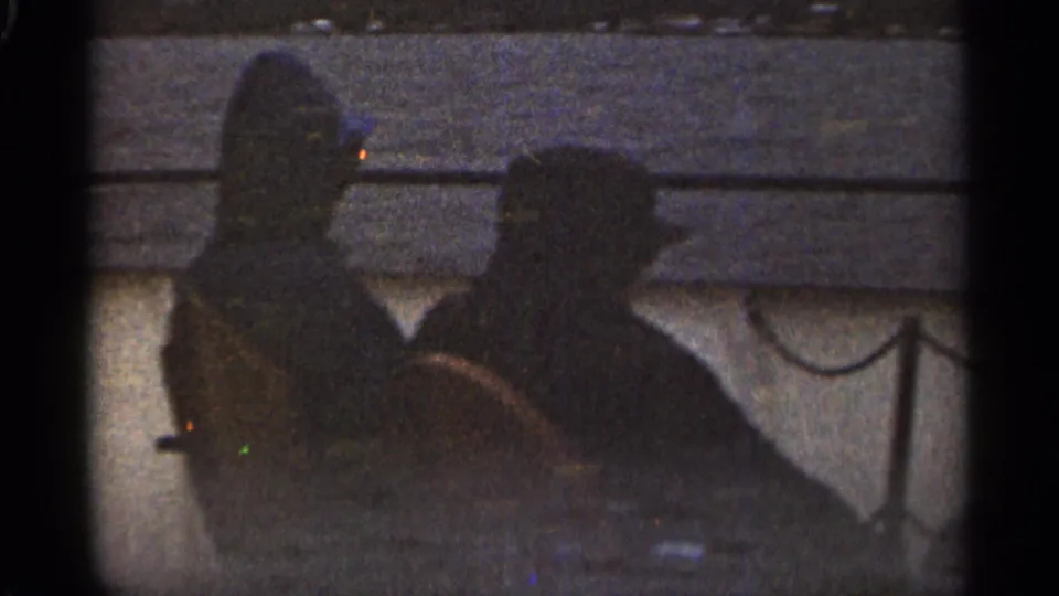 In the 1965 Duluth 8mm clip, two adult men sit close together on a boat, their faces mostly obscured by shadow and film grain.