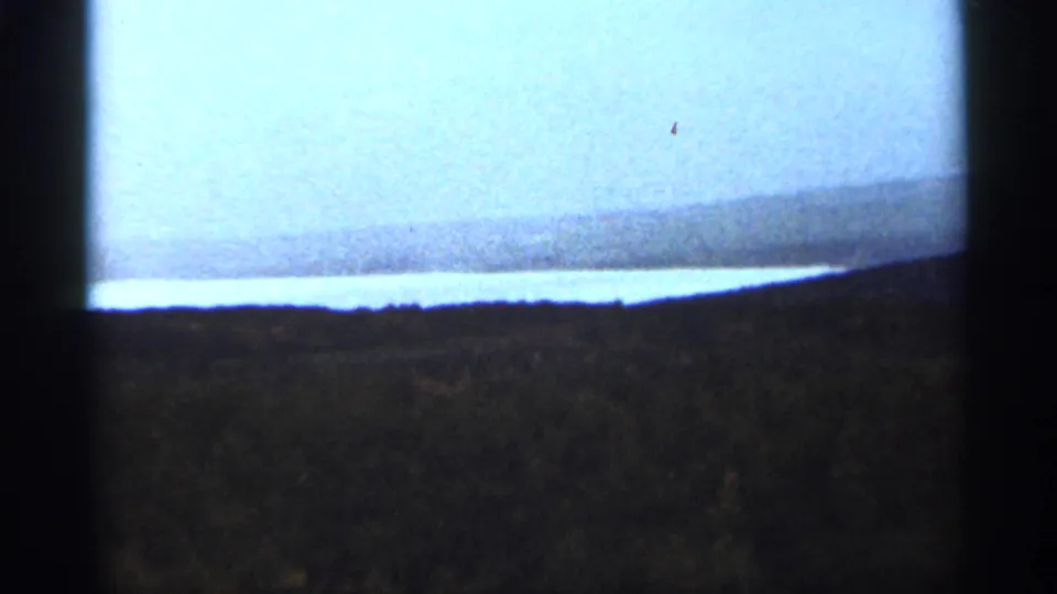1965 8mm view near Duluth showing pale sky, a bright water band, distant low land, and dark foreground terrain.
