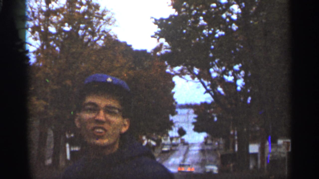 Young man in blue baseball cap and glasses near a tree-lined street.