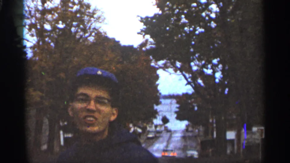 Young man in a blue baseball cap and glasses smiles toward the camera on a Duluth street in 1965 home-movie footage.