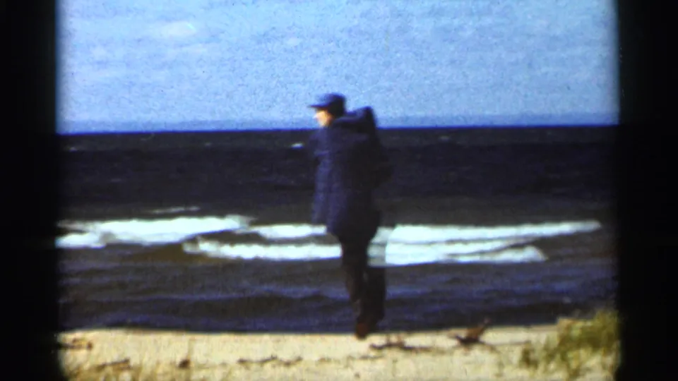 A man in a dark jacket and cap stands near the waterline on a Duluth beach, looking across the waves.