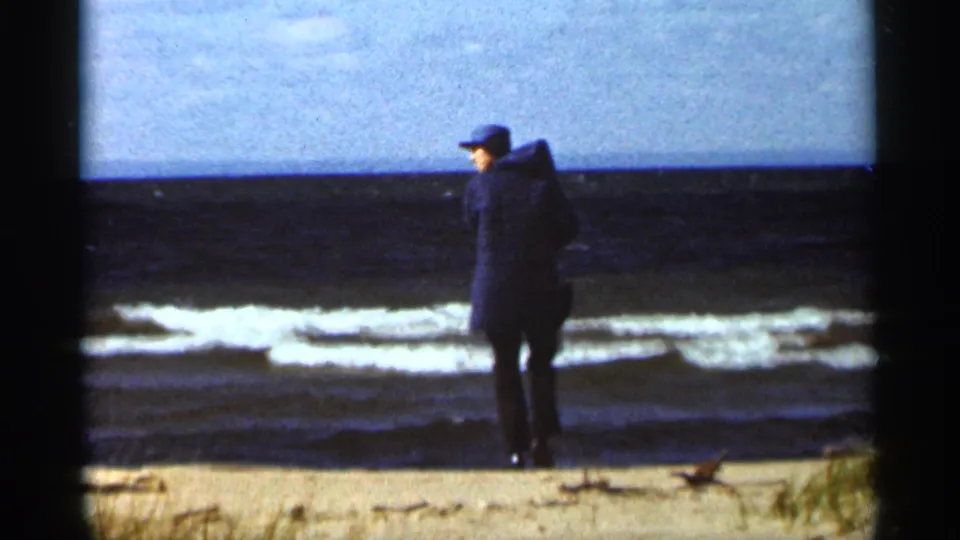 A man in a dark hooded coat stands near the Duluth shoreline in 1965, with waves and pale sky behind him.