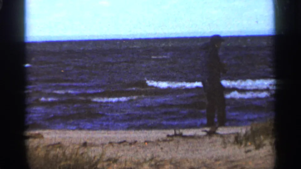 A distant man stands near the Duluth beach waterline in a dark coat, framed by lake waves and a pale horizon.