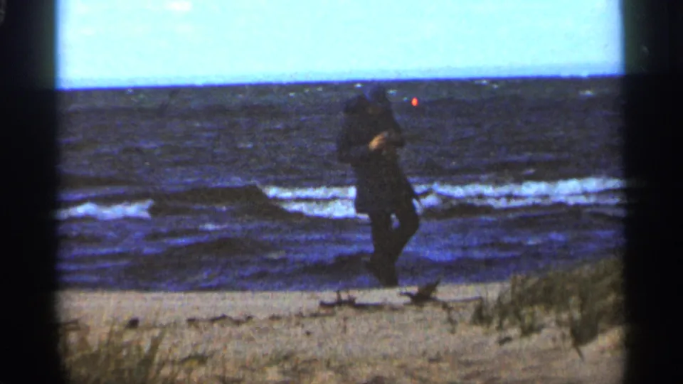 The man walks along the Duluth beach in 1965, carrying or holding something near his side as waves break behind him.