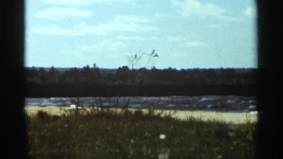 The Duluth lakeshore appears in a wider 1965 home-movie view with grass, waves, and a low wooded horizon.