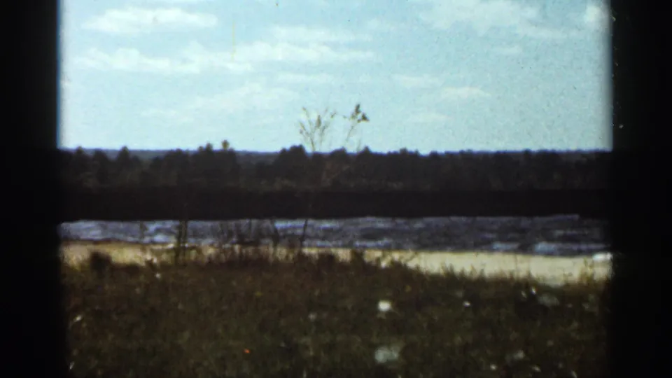 A wide 1965 Duluth shoreline frame shows rough lake water beyond grass and a distant treeline beneath pale clouds.