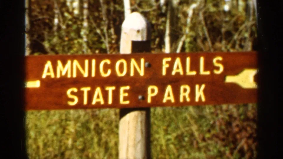 1965 8mm frame of the Amnicon Falls State Park wooden sign, including the park name, white lettering, arrow, signpost, and wooded background.