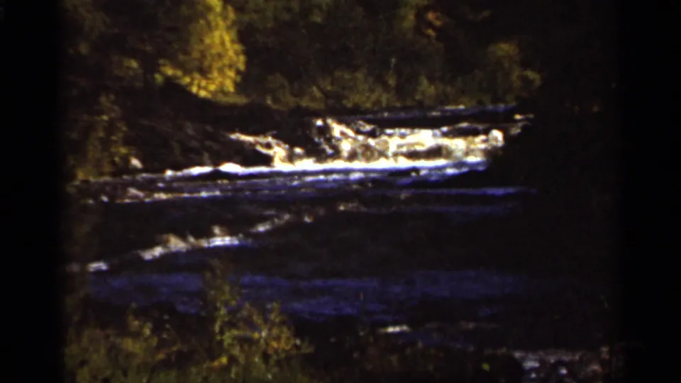 1965 8mm frame of water scudding over rocks at Amnicon Falls, seen in dim natural light.