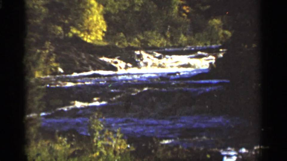 1965 Duluth 8mm frame of white water moving across a rocky river section beneath trees.